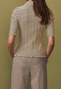 Sweter w paski w kolorze kremowym i czarnym, z krótkimi rękawami o falbaniastych brzegach oraz dopasowanym kroju. Tło jest oliwkowe.