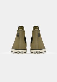 Adidași Converse All Star de culoare verde măslinie, design înalt, țesătură texturată, accente negre, logo proeminent pe toc și talpă albă contrastantă.