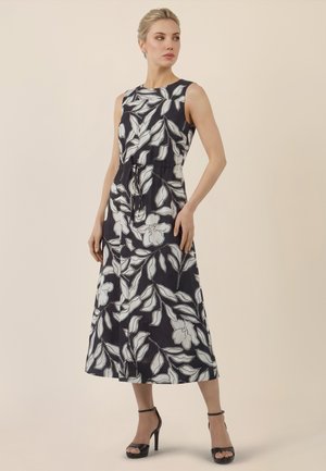 Frau trägt ärmelloses schwarzes Midikleid mit weißem Blumenmuster und schwarze Sandalen mit hohen Absätzen, steht vor einem beigen Hintergrund.