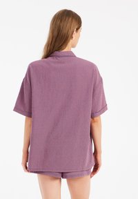 Camicetta a maniche corte, a scacchi nei toni di viola e rosso. Presenta un colletto e una vestibilità comoda. Indossata con pantaloni corti abbinati, in un tessuto leggero.