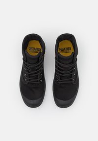 Zapatillas negras de caña alta con parte superior de lona y punta de goma. Presentan cordones negros y una plantilla amarilla con la marca "PALLADIUM".