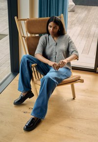 Pilkas megztas trumpomis rankovėmis kardiganas, nuskalbti mėlyni plačiomis kojomis džinsai ir juodi lakinės odos mokasinai, sėdintys ant medinės kėdės prie lango.