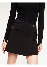 Černá sukňa s obalem vyrobená z hladké tkaniny, obsahující dvě přední kapsy se zlatými knoflíky a čistý lem. Dlouhý rukáv černého topu je částečně viditelný.