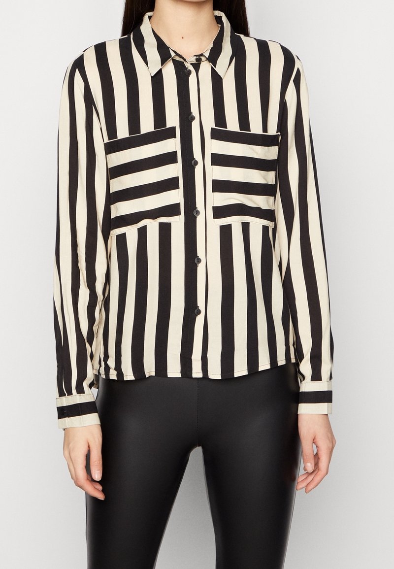 Femme portant une chemise à boutons noire et blanche à rayures verticales avec deux poches poitrine à rayures horizontales et un pantalon noir ajusté.