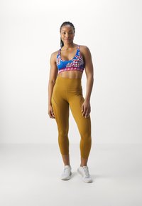 Sujeción deportiva con patrón geométrico azul y ribete rosa, combinada con leggings amarillo mostaza de cintura alta con costuras laterales y logo. Zapatillas blancas.