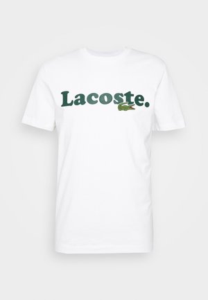 Bílá bavlněná tričko s krátkým rukávem, se zeleným logem "Lacoste" a malou zelenou grafikou krokodýla. Okruglý výstřih.