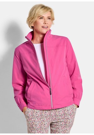Femme blonde portant une veste zippée rose vif et un pantalon à motifs, debout les mains dans les poches, regardant sur le côté et souriant doucement.