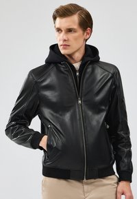 Zwarte leren jas met een rits aan de voorkant, geribde manchetten en een zwarte hoodie eronder. Bevat zijzakken en heeft een strakke, getailleerde pasvorm.