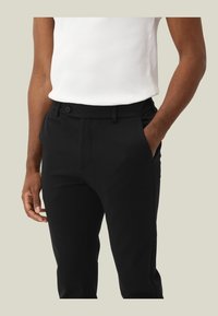 Schwarze Hose mit glatter Textur, schlanker Passform und seitlichen Taschen. Kombiniert mit einem weißen, gerippten Tanktop. Maßgeschneiderter Bund mit Gürtelschlaufen.