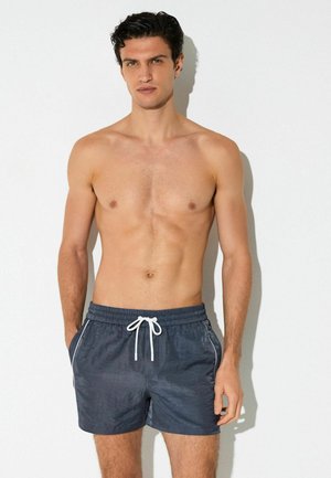 Säratuta mees, kellel on lühikesed tumedad juuksed, kes kannab halli ujumispükse valge nööriga, seisab ühetoonilise hele tausta ees.