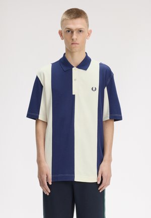 Jeune homme aux cheveux courts portant un polo à rayures verticales bleu marine et blanc, se tenant devant un arrière-plan clair uni.