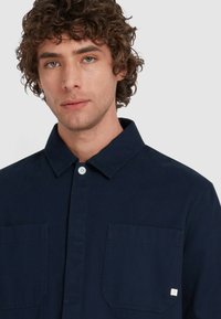 Mężczyzna z kręconymi włosami, ubrany w ciemnoniebieską koszulę na guziki z kieszonkami na piersi, patrzy prosto przed siebie na jasnym, jednolitym tle.