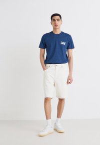T-shirt en coton bleu marine avec le logo "Lee" associé à un short en denim blanc. Le mannequin porte des sneakers blanches et des chaussettes. Design minimaliste, couleurs neutres.