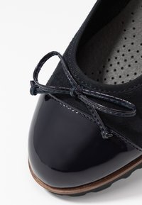 Detal czarnych lakierowanych baletków z błyszczącą kokardą i perforowaną wyściółką.