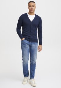Mørkeblå strikket cardigan med V-hals, knappelukking og ribbestrikket tekstur på skuldrene, kombinert med lyseblå jeans og hvite sneakers.