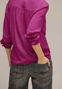 Blusa fucsia con vestibilità ampia, maniche lunghe arrotolate; abbinata a jeans grigio scuro con tasche posteriori ricamate uniche.