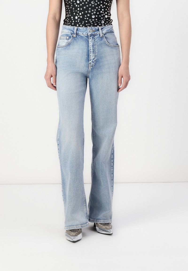 Light blue flared jeans with a smooth texture, featuring five pockets, a zipper fly, and a classic denim design, paired with metallic shoes.