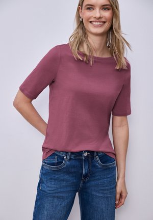 Lächelnde Frau, die ein kurzärmeliges Shirt in Mauve und blaue Jeans trägt, steht mit einer Hand hinter dem Rücken vor einem schlichten hellen Hintergrund.