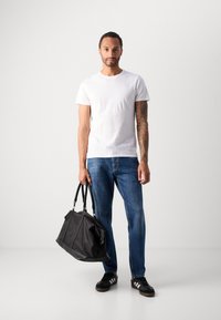 Geantă de voiaj din piele neagră cu două mânere, tricou alb simplu, blugi, și adidași negri cu accente albe. Modelul stă în picioare.