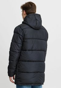 Chaqueta acolchada negra con capucha, con secciones acolchadas y un corte relajado. La tela parece suave; el diseño es minimalista y práctico.