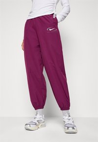 Person som bär lila Nike sportbyxor med elastiska mudd och vita och grå Nike sneakers, stående på ett vitt golv.