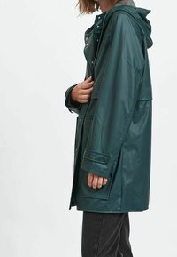 Grön vattentät regnrock med huva, tryckknappslåstningar, sidfickor och en avslappnad passform. Strukturerad tyg med slät finish.