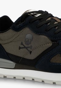 Zapatilla de deporte negra y verde oscuro con cordones negros, que presenta un logo de calavera y huesos cruzados en el lateral y una suela texturizada blanca.