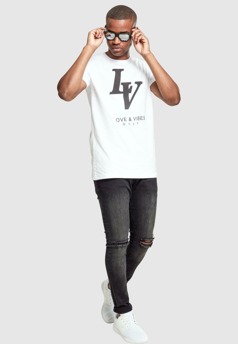 Tricou alb cu logo-ul negru "LV" și textul "LOVE & VIBES ONLY"; asortat cu blugi negri distruși și teniși albi.