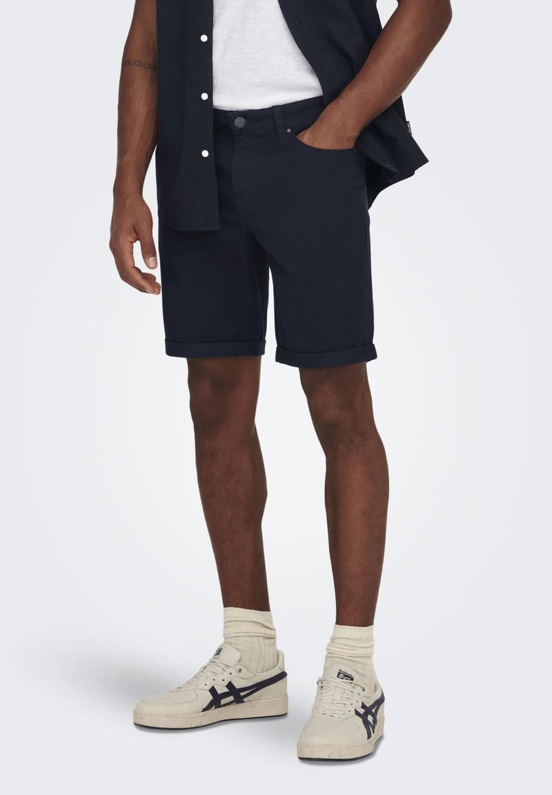 Marineblå rullesort med knaplukning, sidelommer og en afslappet pasform, parret med hvide sneakers og lette ankelsokker.