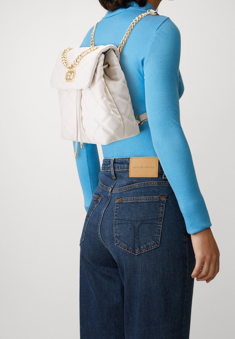 Quilted white backpack with a chain strap, featuring a fold-over top and zip closure. Worn on a blue sweater with blue denim jeans.
