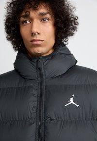 Čierna prešívaná bunda s vysokým golierom a predným zipsom. Na ľavej hrudi sa nachádza biely logo Jumpman a má hladkú, lesklú textúru.