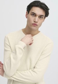 Maglione a coste di un beige chiaro, con scollatura a V e maniche lunghe. La texture è morbida e leggermente strutturata, progettata per un fit rilassato.
