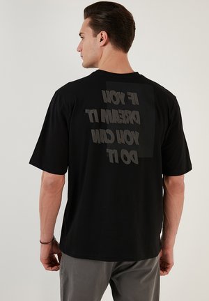 Camiseta de algodón negra con un gran diseño de texto en espejo en la parte posterior: "SI LO SUEÑAS, LO PUEDES HACER." Mangas cortas y corte relajado.