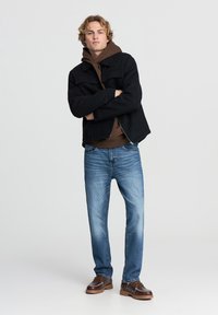 Giacca nera con zip, felpa marrone, jeans azzurri chiari e scarpe marroni allacciate. Modello in piedi con le braccia incrociate, sfondo neutro.
