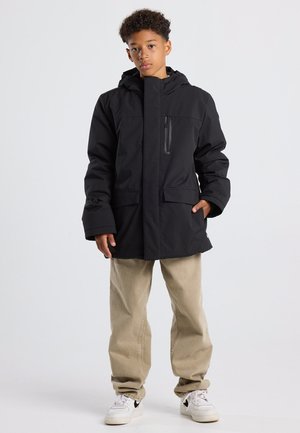 Junge trägt eine schwarze Winterjacke mit Kapuze, beige Hosen und weiße Sneakers, steht mit einer Hand in der Tasche vor einem schlichten hellen Hintergrund.
