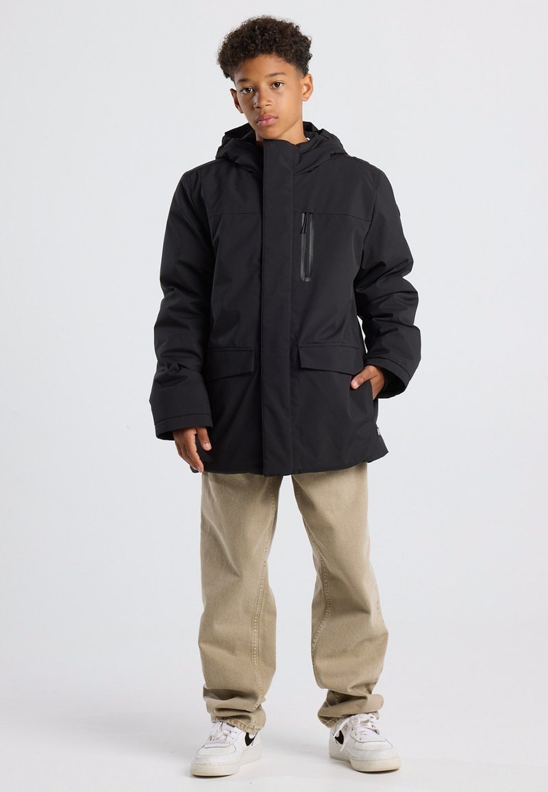 Poika, jolla on päällä musta hupparillinen talvitakki, beige housut ja valkoiset tennarit, seisoo yksi käsi taskussa vaaleaa taustaa vasten.