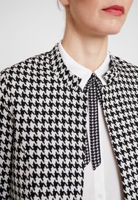 Chaqueta de cuadros houndstooth en blanco y negro con cuello redondo, combinada con una camisa blanca abotonada que presenta un detalle de corbata negra.
