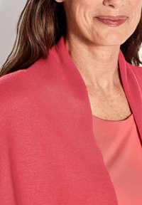 Châle en tricot rose drapé sur un haut rose plus clair. Le tissu a une texture subtile, avec une apparence lisse et douce et un design à l'avant ouvert.