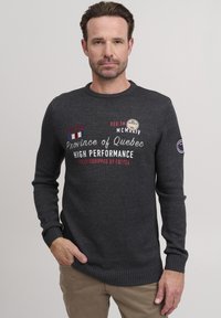 Grå stickad tröja med rund halsringning, prydd med röd och vit text och lappar, ribbade muddar och en strukturert yta.