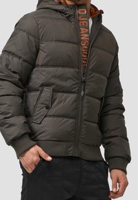 Homme portant une veste matelassée gris foncé avec des poches zippées et à boutons-pression, une main dans la poche d'un pantalon noir, doublure intérieure de marque visible.