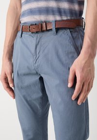 Hellblaue Baumwoll-Chinohose mit glatter Textur, kombiniert mit einem braunen Ledergürtel mit silberner Schnalle, Hände ruhen auf der Taille.