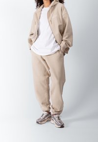 Sudadera beige con cierre de cremallera y logo, combinada con pantalones de chándal a juego. El modelo lleva una camiseta blanca y zapatillas grises. Textura de tela suave.