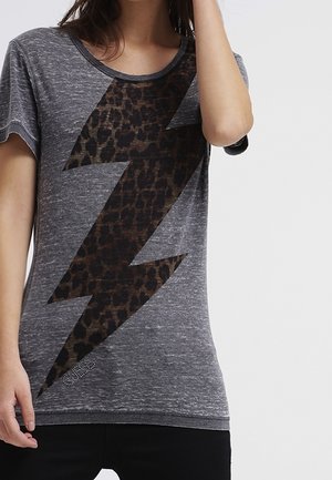 Femme portant un t-shirt gris avec un grand motif de foudre en léopard sur le devant, levant une main près de son visage.