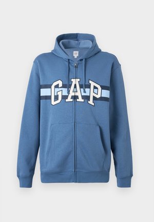Blå hettegenser med glidelås, frontlommer og hvite og marineblå striper med stor "GAP"-tekst over brystet.
