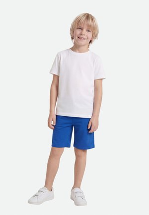 Ξανθό αγόρι που στέκεται και χαμογελά, φορώντας ένα απλό λευκό t-shirt, μπλε σορτς και λευκά αθλητικά παπούτσια με βέλκρο σε λευκό φόντο.