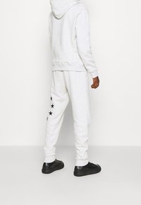 Ljusgrå hoodie och matchande mjukisbyxor med ett svart stjärnmönster längs vänster ben. Svarta sneakers syns längst ner.