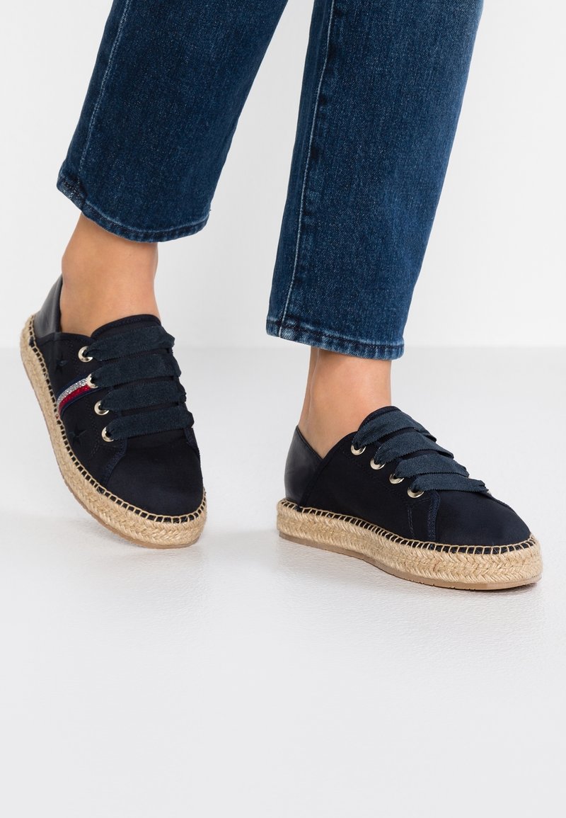 Baskets espadrilles bleu marine avec larges lacets, semelle en jute tissée et détail latéral rouge et blanc portées avec un jean bleu.