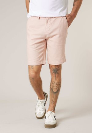 Homme portant un short rose clair, des baskets blanches et une chemise blanche, avec des tatouages visibles sur les deux jambes, marchant sur un fond neutre.
