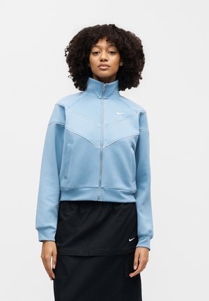 Kvinde med krøllet hår iført en lyseblå lynlåstrøje og en sort nederdel med et lille hvidt Nike-logo på hvert tøjstykke.