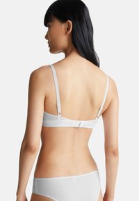 Žena s dlouhými černými vlasy, oblečená v bílém podprsenkovém a kalhotkovém kompletu, zachycena zezadu na jednoduchém světlem pozadí.